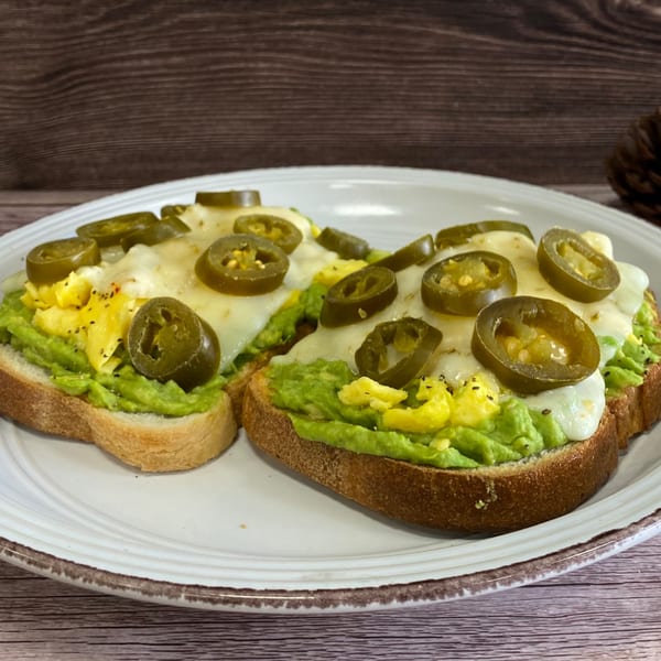 Two slices of avocado toast topped with scrambled eggs, melted cheese, and sliced jalapeños.