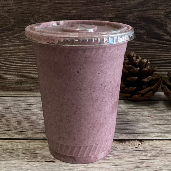 A purple smoothie in a clear plastic cup with a lid, on a rustic wooden surface with pinecones.