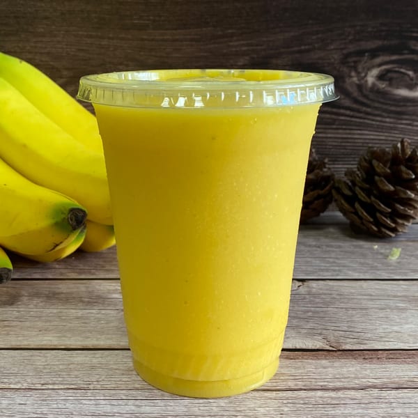 A bright yellow banana smoothie in a clear cup, with bananas and pinecones on a wooden table.