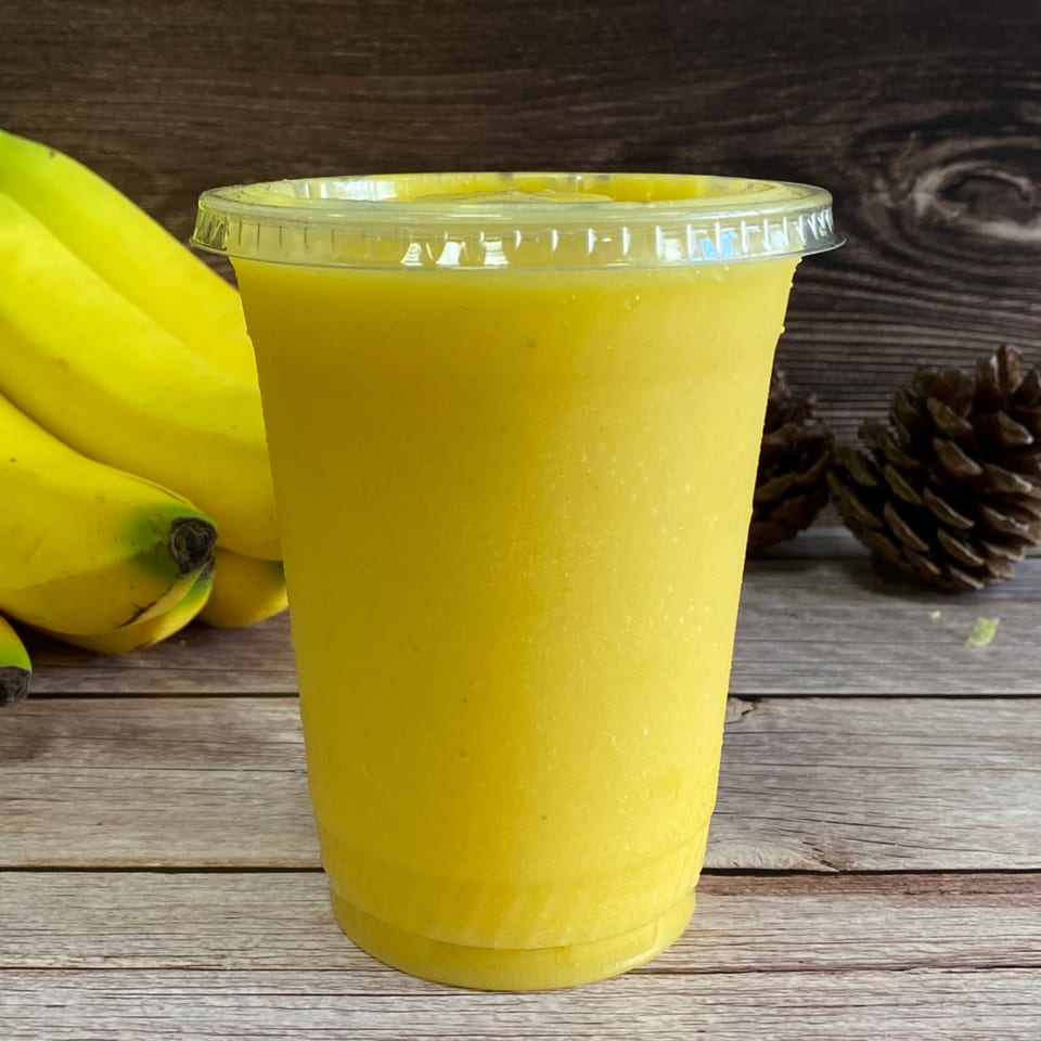 A bright yellow banana smoothie in a clear cup, with bananas and pinecones on a wooden table.