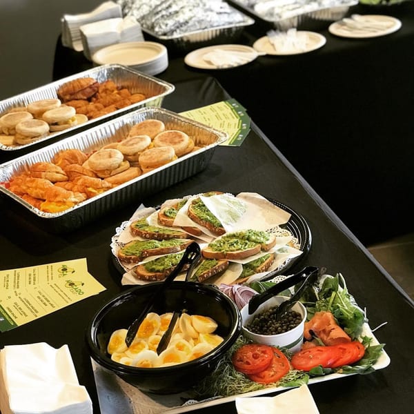 A catering spread of Avocado Toast, breakfast sandwiches, hard-boiled eggs, tomatoes, and capers on a black tablecloth.