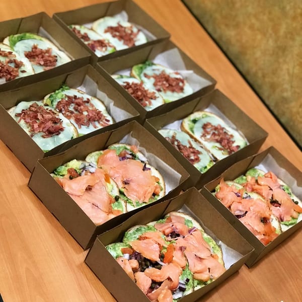 Rows of Avocado Toast with smoked salmon and bacon in individual brown takeout boxes.