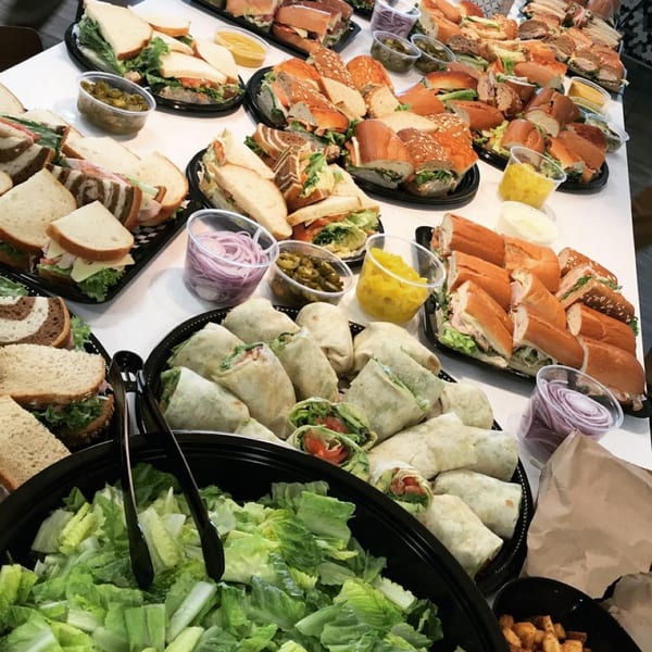 A catering spread of various sandwiches, wraps, a large bowl of salad, and condiments.