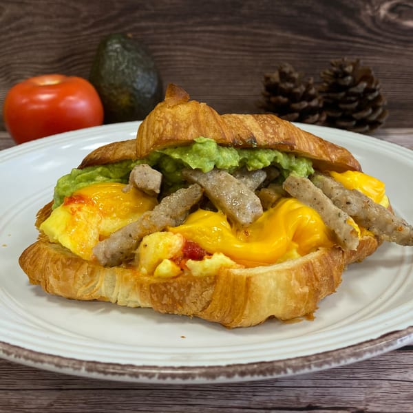 Croissant filled with scrambled eggs, melted cheese, sausage, and guacamole on a white plate.