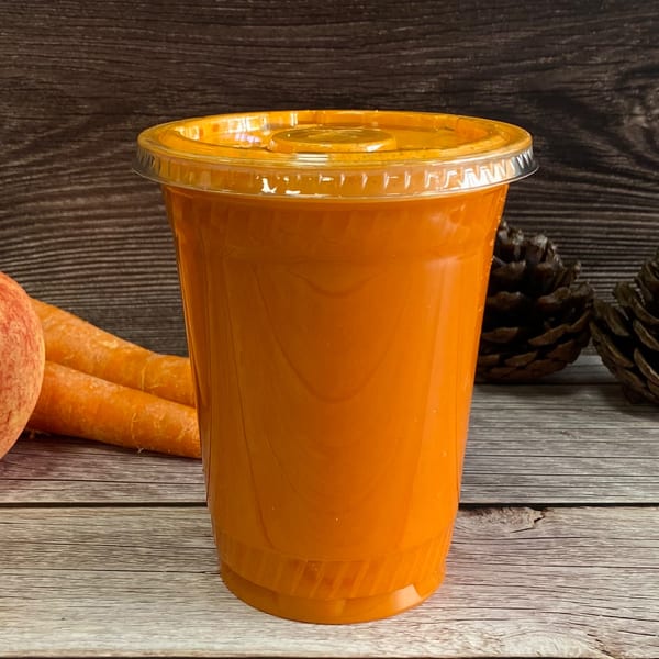 Freshly squeezed orange juice in a clear plastic cup with carrots and pinecones on a wooden table.